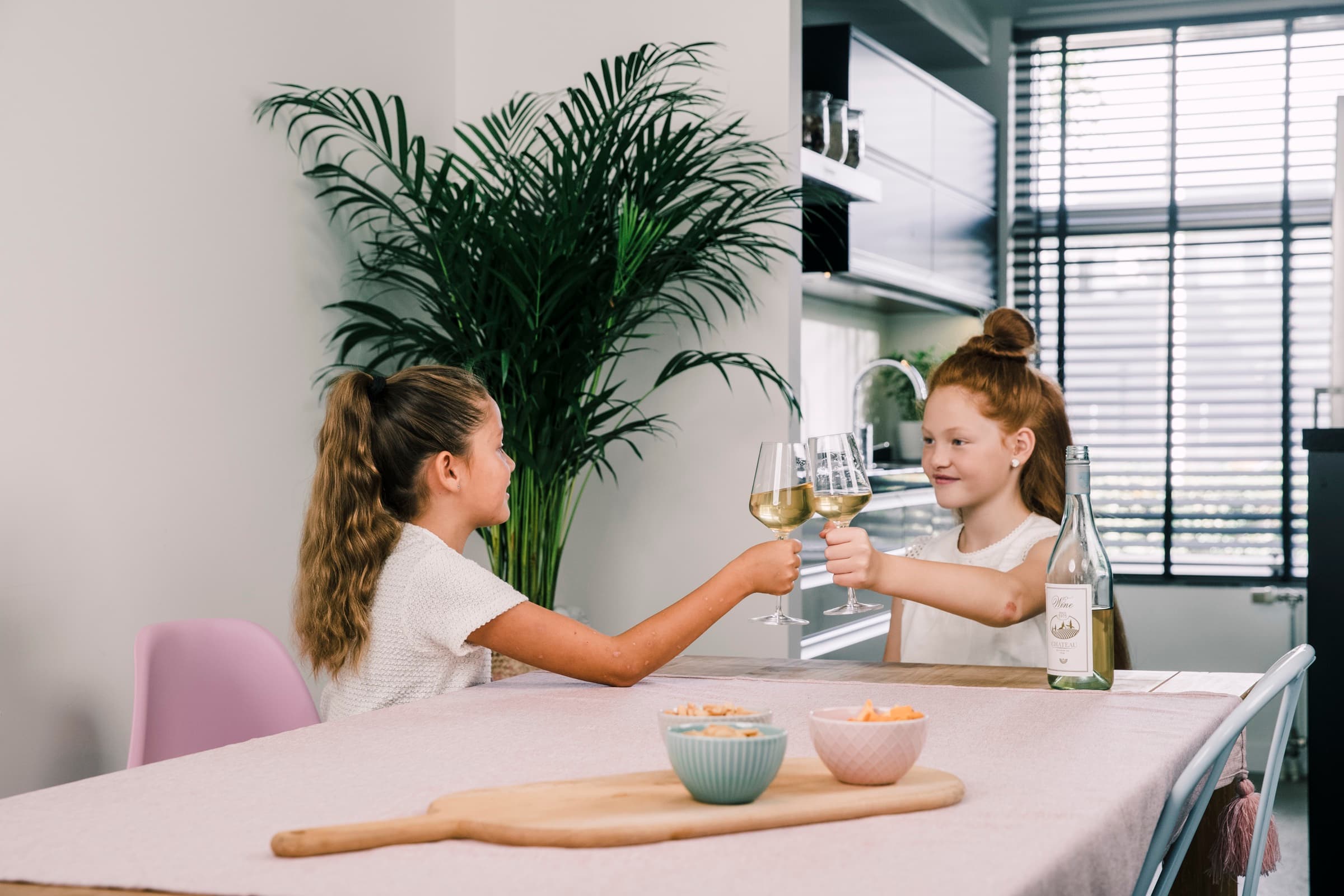 Twee meisjes aan dezelfde keukentafel proosten met een wijnglas, precies zoals de vrouwen.