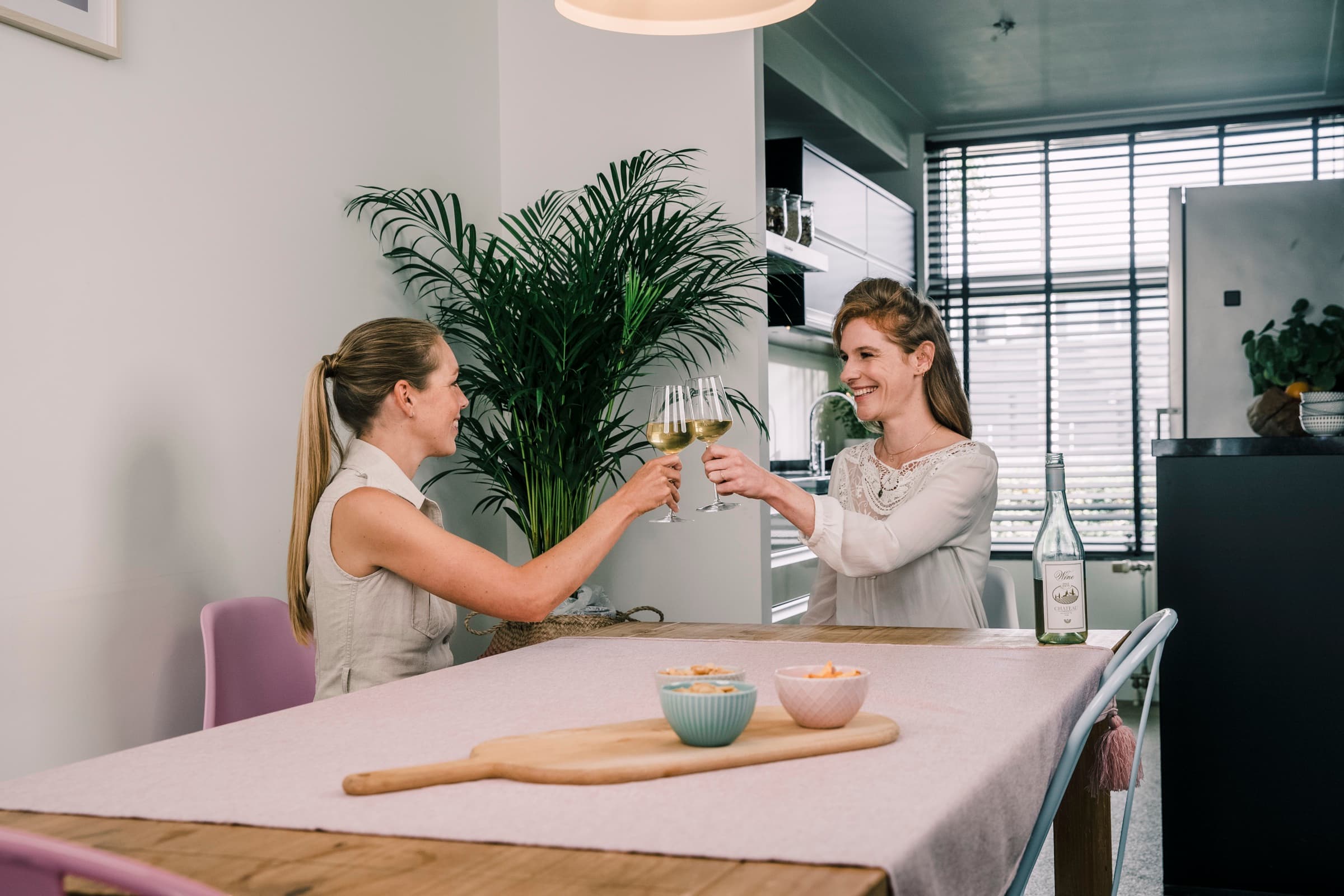 Twee vrouwen aan de keukentafel proosten met een glas witte wijn.